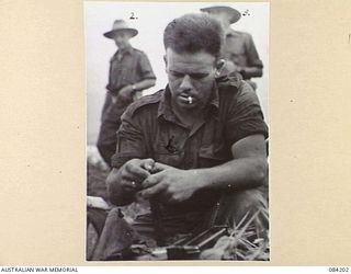 SUAIN PLANTATION, NEW GUINEA. 1944-12-10. LANCE-BOMBARDIER K. D. L. MCCREDIE, 2/6 SURVEY BATTERY, (1), FILLING AN OWEN GUN MAGAZINE BEFORE THE COMMENCEMENT OF A SURVEY TRIP. MAJOR-GENERAL J. E. S. ..