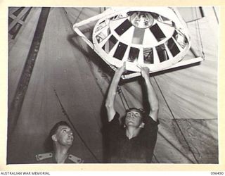 RABAUL, NEW BRITAIN, 1945-09-12. MAJOR W.G. MILLER (1) AND PRIVATE SMITH (2), 4 FIELD AMBULANCE, ADJUSTING AN OPERATING THEATRE LAMP IMPROVISED FROM GALVANISED IRON, BISCUIT TINS AND CANTEEN ..