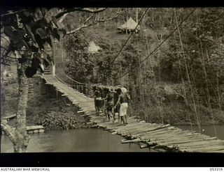 Sogeri, New Guinea. 1943-06-26. Native labourers carrying a log across the Laloki River suspension bridge at the New Guinea School of Signals. The labourers were supplied by the Australian and New ..