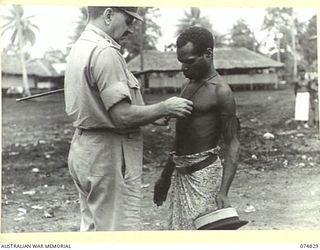 LAE, NEW GUINEA. 1944-07-22. MAJOR GENERAL B.M. MORRIS, DSO, GENERAL OFFICER COMMANDING, AUSTRALIAN NEW GUINEA ADMINISTRATIVE UNIT, PLACING A MEDAL AROUND THE NECK OF GARTER, A NATIVE "DOCTOR" WHO ..