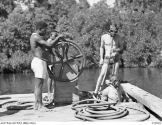 NX170988 Spr R Skidmore (standing), and NX79944 Sapper (Spr ) E Nichols, both of the 17th Field Company, have their native boys mann the pumps of their improvised diving gear as they move into the ..