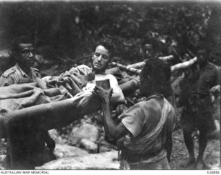 Native stretcher bearers stop at a river to give a drink of water to their patient, Private A. Baldwin, of the 2/33rd Battalion