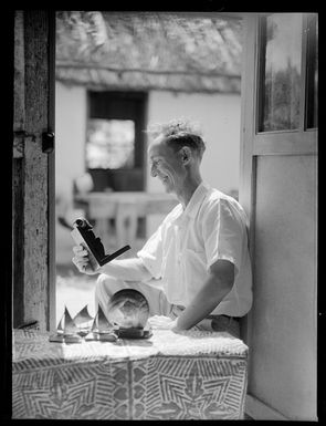 Unidentified European man sitting in a doorway of a house holding a carving of Tangaroa, a Cook Island god, includes other shell and carvings of boats on the table, Rarotonga, Cook Islands