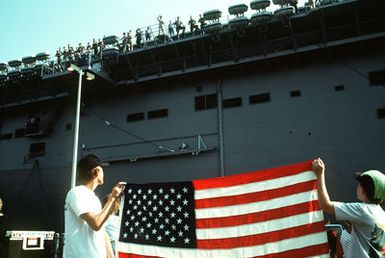 Two boys hold up an American flag as the amphibious assault ship USS GUAM (LPH-9) departs for the Persian Gulf in response to Iraq's invasion of Kuwait