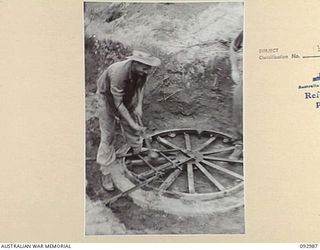 WEWAK AREA, NEW GUINEA, 1945-06-07. CORPORAL E.J. PAYNE, 2/3 FIELD REGIMENT, ROYAL AUSTRALIAN ARTILLERY, HAMMERING THE LAST STEEL PEG INTO THE GROUND TO HOLD THE BASE PLATE OF THE GUN IN POSITION, ..