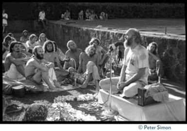 Ram Dass seated on a small platform, arms raised, during his appearance at Andrews Amphitheater, University of Hawaii