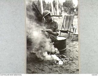 NEW GUINEA, 1942-08. ONE WAY TO COMBAT THE MOSQUITO MENACE IN NEW GUINEA. THIS AUSTRALIAN SOLDIER READS MAIL FROM HOME OVER A FIRE OF SMOKING DRIED COCONUT SHELLS, WHICH GIVES OFF A VERY PUNGENT ..