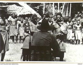 WIDERU, NEW GUINEA, 1943-10-21. NX155085 CAPTAIN R.G. ORMSBY OF THE AUSTRALIAN AND NEW GUINEA ADMINISTRATIVE UNIT SITTING AT HIS DESK, ADDRESSING THE VILLAGE NATIVES IN A TALK ON THE WAR AND ..