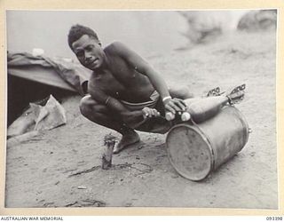 BERRY'S HILL, CENTRAL BOUGAINVILLE, 1945-06-27. A NATIVE CARRIER TYING TWO 4.2 INCH MORTAR BOMBS TO A TIN OF TEA DURING THE MOVEMENT OF SUPPLIES TO 7 INFANTRY BATTALION TROOPS ALONG THE NUMA NUMA ..