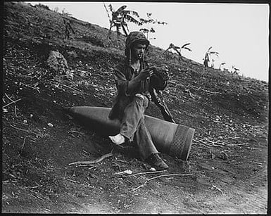 Using an unexploded 16-inch naval shell for a resting place, Marine Private First Class Raymond Hubert, shakes a three-day accumulation of sand from his boondocker. Saipan, July 4, 1944.