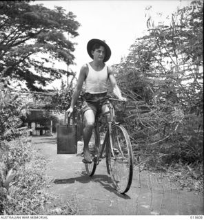 1942-11-19. NEW GUINEA. KOKODA. PRIVATE R.S. BRETT (N.S.W.) COOKS FOR SOME OF THE LADS IN KOKODA. HE RIDES A BROKEN DOWN JAPANESE BICYCLE. IT HAS NO TYRES AND ONLY HALF A HANDLE-BAR. (NEGATIVE BY ..