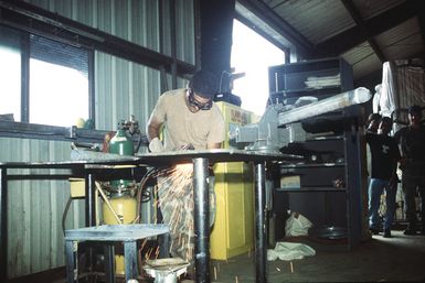 Steelworker Second Class (SW2) Frank Luevanos uses a cutting torch to cut a piece of sheet metal in the welding shop of the Naval Mobil Construction Battalion Three (NMCB-3) Civic Action Team compound