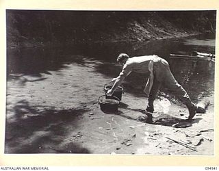 PARCHY RIVER, NEW GUINEA, 1945-07-23. SAPPER A.D. LEE, 10 PLATOON, 2/14 FIELD COMPANY, ROYAL AUSTRALIAN ENGINEERS, COOK AT THE ENGINEER'S CAMP, PLACING TWO DIXIES CONTAINING JELLY AND CUSTARD INTO ..