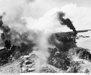 KAVIENG, NEW IRELAND. ? 1944-03. AERIAL PHOTOGRAPH OF BOMBS BURSTING AT THE MAIN JAPANESE BASE. A FLOATPLANE IS BLAZING IN WATER BEYOND THE TOWN