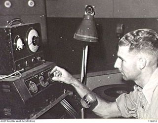 LAE, NEW GUINEA. 1945. SERGEANT P. DENNIS, ANNOUNCER, AT THE ANNOUNCER'S CONTROL PANEL OF RADIO LAE, A 50-KILOWATT RADIO TRANSMITTER WHICH HAS BEEN MADE FROM SALVAGED ALLIED AND ENEMY WIRELESS ..