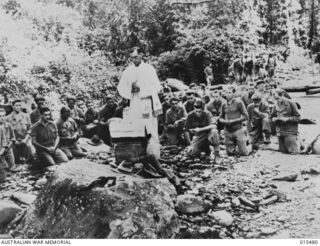 1943-08-10. NEW GUINEA. MOUNT TAMBU FIGHTING. WITHIN SOUND OF RIFLE, ARTILLERY AND MORTAR FIRE, FATHER THEO O'KEEFE, OF ST. PATRICK'S CHURCH, CAMPERDOWN, VIC, HOLDS MASS ALONGSIDE A MOUNTAIN STREAM ..