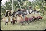 Dancers in grass skirts and matching headress; some with western dress or tops, and white handkerchiefs tied around head or arm