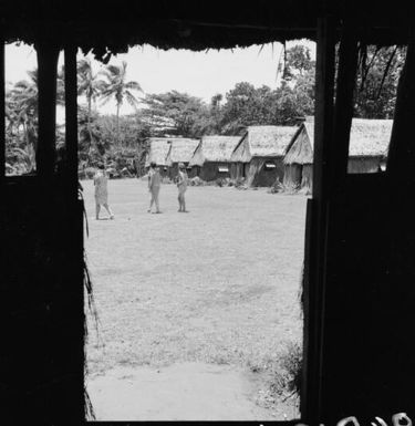 Three women walking in front of a row of grass huts, Nasalai, Fiji, 1966 / Michael Terry